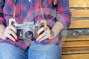 Girl in blue jeans and checkered shirt is sitting on the wooden bench and holding an old film camera in her hands.