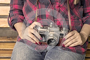 Girl in blue jeans and checkered shirt is sitting on the wooden bench and holding an old film camera in her hands.