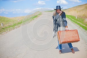 A girl in blue jeans, a poncho, a black leather hat with an old brown suitcase in her hands is on the road.