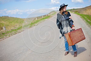 A girl in blue jeans, a poncho, a black leather hat with an old brown suitcase in her hands is on the road.