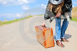 A girl in blue jeans, a poncho, a black leather hat with an old brown suitcase in her hands is on the road.