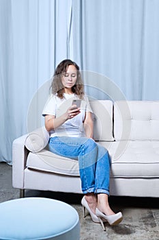 A girl in blue jeans and a white t-shirt looks at the phone while sitting on a light-colored sofa
