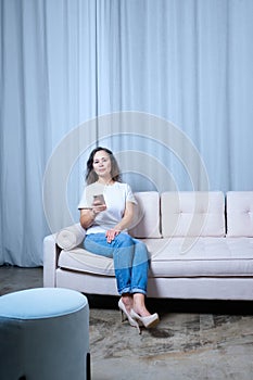 a girl in blue jeans and a white t-shirt looks at the phone while sitting on a light-colored sofa