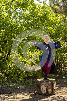 A woman in a blue kimono poses with a sword in the forest standing on a stump