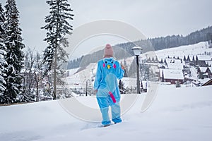 Girl in blue, pink unicorn pijama kigurumi outdoor in front of the wood houses on the ski report in snow mountains.
