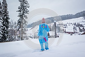 Girl in blue, pink unicorn pijama kigurumi outdoor in front of the wood houses on the ski report in snow mountains. photo