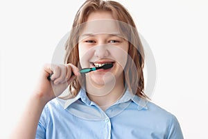 A girl in a blue shirt with blonde hair brushes her teeth and smiles against a light background, looking at the camera