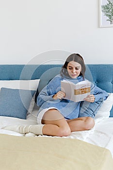 Girl in a blue sweater reading a book on psychology sitting on the bed in a cozy interior