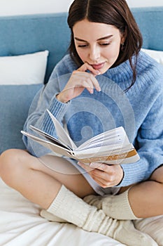 Girl in a blue sweater reading a book on psychology sitting on the bed in a cozy interior