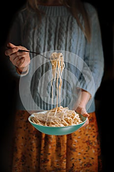 A girl in a blue sweater and yellow dress holds a bowl of spaghetti.