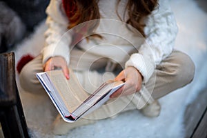 A girl with a book is sitting on the floor on a furry white carpet. Dressed in a white sweater and light trousers with a