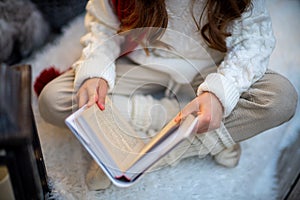 A girl with a book is sitting on the floor on a furry white carpet. Dressed in a white sweater and light trousers with a