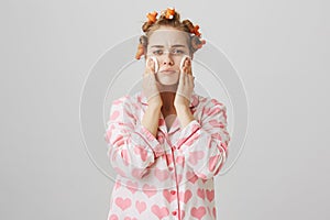 Girl is bothered with new acne. Portrait of gloomy good-looking young woman in hair curlers and pyjamas holding cotton