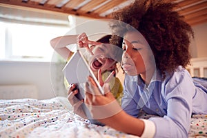 Girl And Boy In Bedroom Lying On Bed Using Digital Tablet And Pulling Funny Faces Together