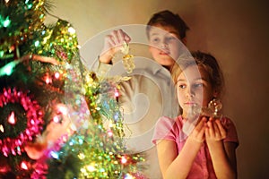 Girl and boy decorated Christmas tree by glass toys at evening.
