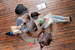 Girl and boy reading books leaning on each other on wooden floor