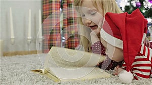 Girl and boy in a Santa hat reading a book in front of the Christmas tree.