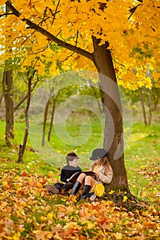 Girl and boy sit under a tree and read a book together in autumn sunny park, children sit in leaves