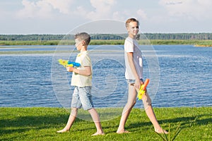 Girl and boy stand on green grass, their backs to each other and play with water pistols, against the blue sky and lake