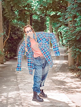 A girl with two braids in a plaid shirt with oversleeves and oversized jeans stands in a pose in a park on a green alley