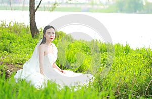 Girl bride in wedding dress with elegant hairstyle, with white wedding dress Sitting in the grass by the river