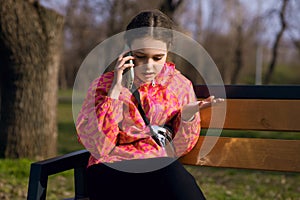 A girl in a bright jacket sits on a park bench and talks on the phone