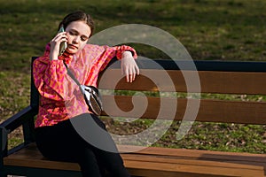 A girl in a bright jacket sits on a park bench and talks on the phone