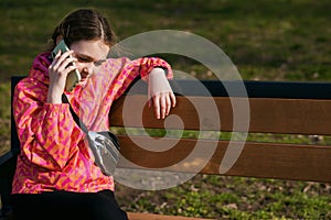 A girl in a bright jacket sits on a park bench and talks on the phone