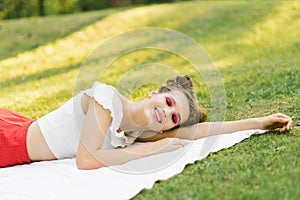 Girl with bright makeup is lying on a spruce blanket on the grass with her eyes closed and smiling. Professional summer makeup in