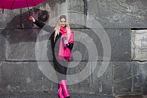 Girl in a bright pink scarf, rubber boots and umbrella on the background of gray stone wall