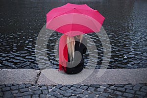 Girl in a bright pink scarf, rubber boots and umbrella sitting on the banks of the river, back. Grey grim landscape.