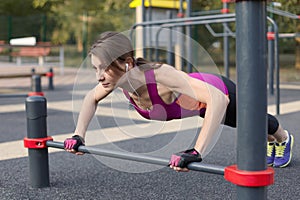 The girl in a bright sportswear pushes up in outdoor sportground. White earphones, protective gloves. Summer, spring, early autumn