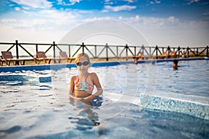 A girl in a bright swimsuit and swimming goggles poses near the pool sunbathing under the summer sun