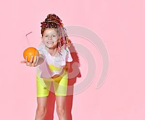 Girl in bright yellow shorts and white t-shirt leaning forward proposing spectator cocktail in a fresh orange, isolated on pink