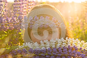 a girl with brown hair and freckles looking like a mermaid playfully looks through the flowers of lupines.