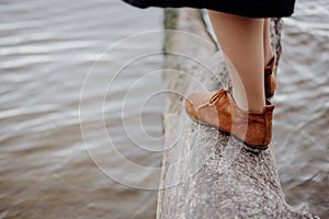 A girl in brown leather boots is standing on a log on the seashore, waves with white foam are rolling on the shore against the