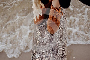 A girl in brown leather boots is standing on a log on the seashore, waves with white foam are rolling on the shore against the