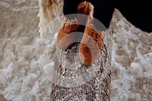 A girl in brown leather boots is standing on a log on the seashore, waves with white foam are rolling on the shore against the