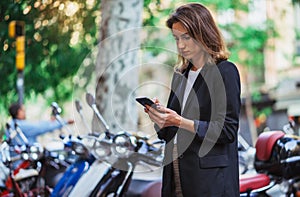 Girl in business clothes holds smartphone against  background mopeds in city summer street outdoors, activ tourist uses a mobile