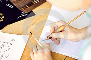 A girl calligrapher paints pen on white paper behind her working wooden table.