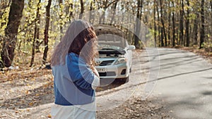 The girl calls the repair service near the broken car on the road in the forest.