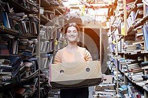 Girl carries a box of books in the library, a portrait of a bookseller against a background of a bookstore