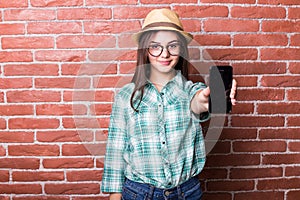 Girl in casual clothes, hat and eyeglasses posing, smiling and demonstrate on smartphone