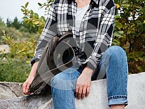 A girl in casual comfortable clothes is sitting on the rocks. A girl in a plaid shirt, blue jeans with a leather backpack, a