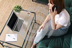 Girl in casual sitting on the sofa using laptop and talking on mobile. Copy space