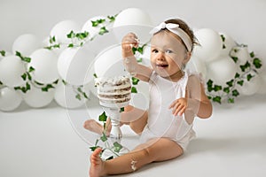 A girl celebrates her first birthday and eats a birthday cake. Decorations for a child`s birthday made of balloons and ivy