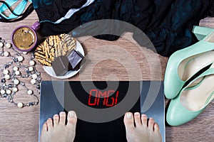 Girl checks her weight on digital scales. Sign omg! on balance surrounded by cup of coffee, sweets and female clothes and shoes photo