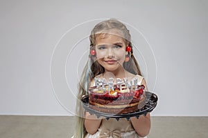 Girl with cherry on ears holds birthday cake with candles, white background, birthday celebration