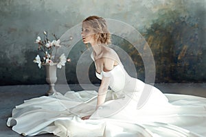 Girl in a chic long dress sitting on the floor. White wedding dress on the bride`s body. Beautiful light dress with a long hem, a