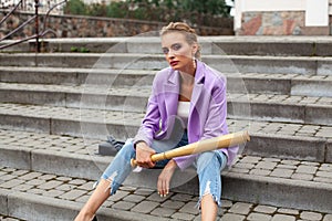 A girl with chic makeup and a stylish hairstyle, sits on the stairs with a baseball bat